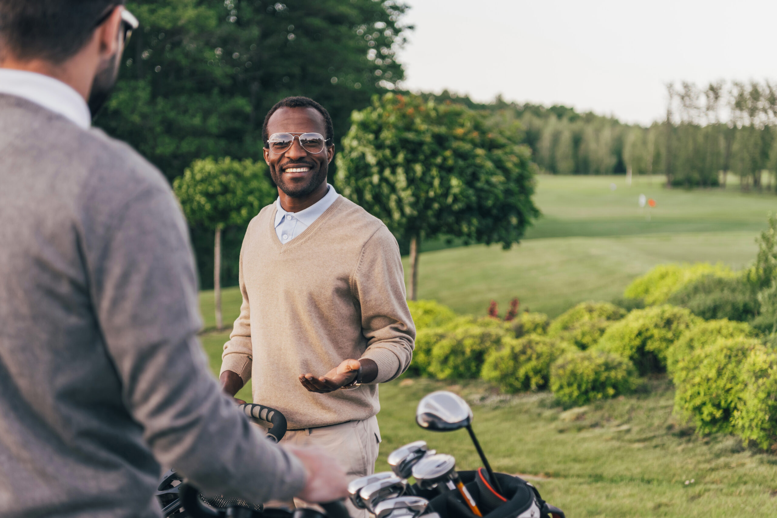 Two smiling men in sunglasses holding golf clubs in bags and talking outdoors